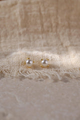 A pair of small gold stud earrings with white pearl-like beads, resting on a textured beige fabric background.
