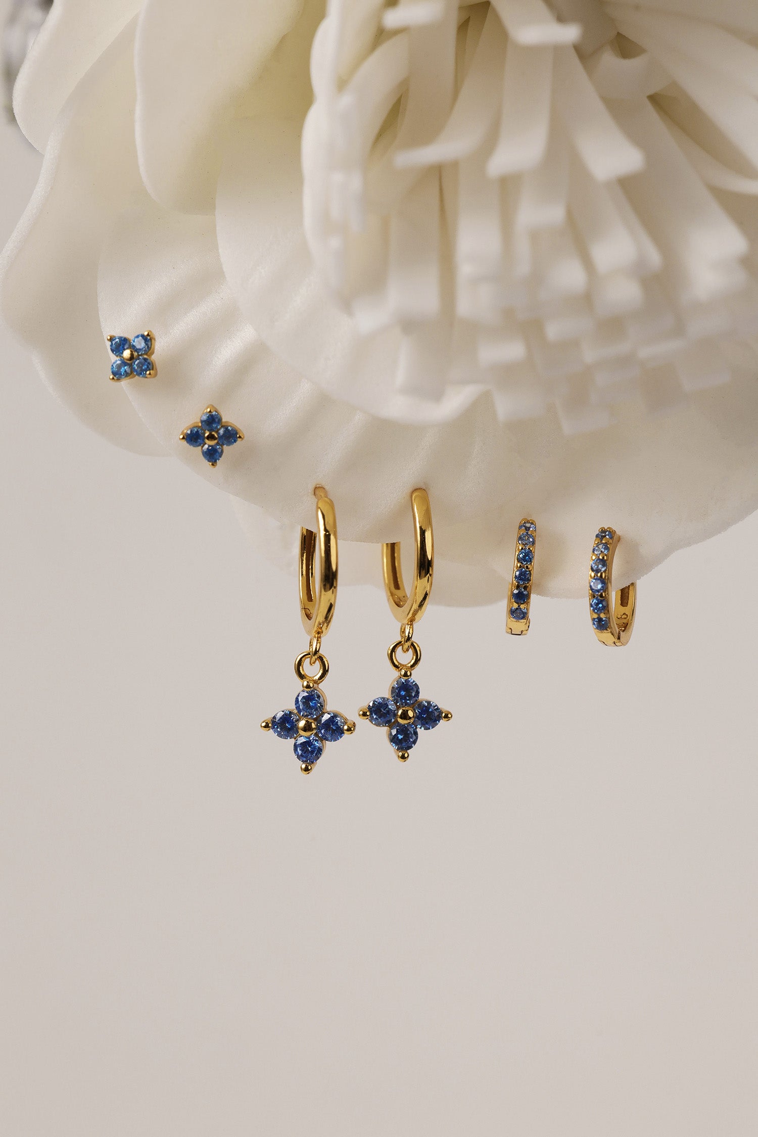 A set of gold-toned hoop and stud earrings with blue crystal floral motifs, displayed against a large white flower backdrop.