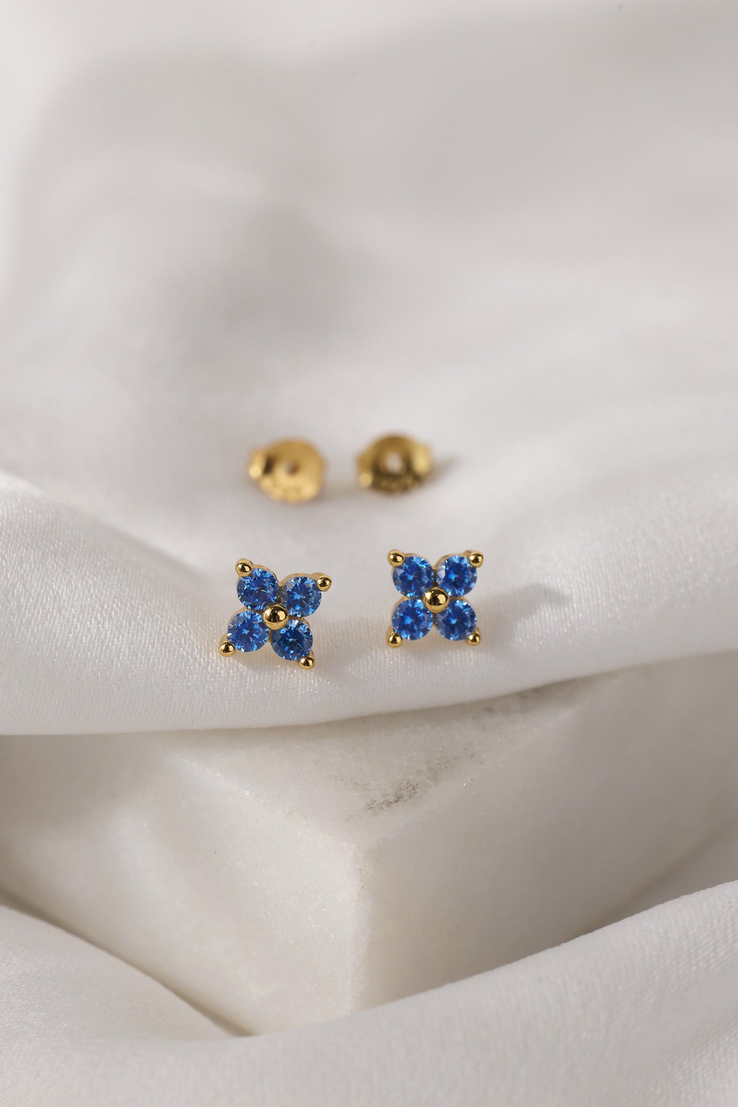 A pair of small gold stud earrings with four blue gemstones arranged in a flower shape, displayed on a soft white fabric surface.