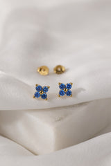A pair of small gold stud earrings with four blue gemstones arranged in a flower shape, displayed on a soft white fabric surface.
