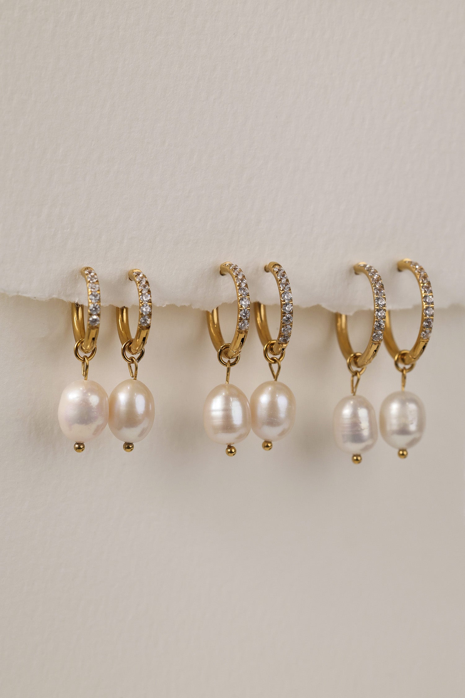 Three sizes of gold pavé waterproof hoop earrings with natural freshwater pearl drops displayed on a textured neutral background