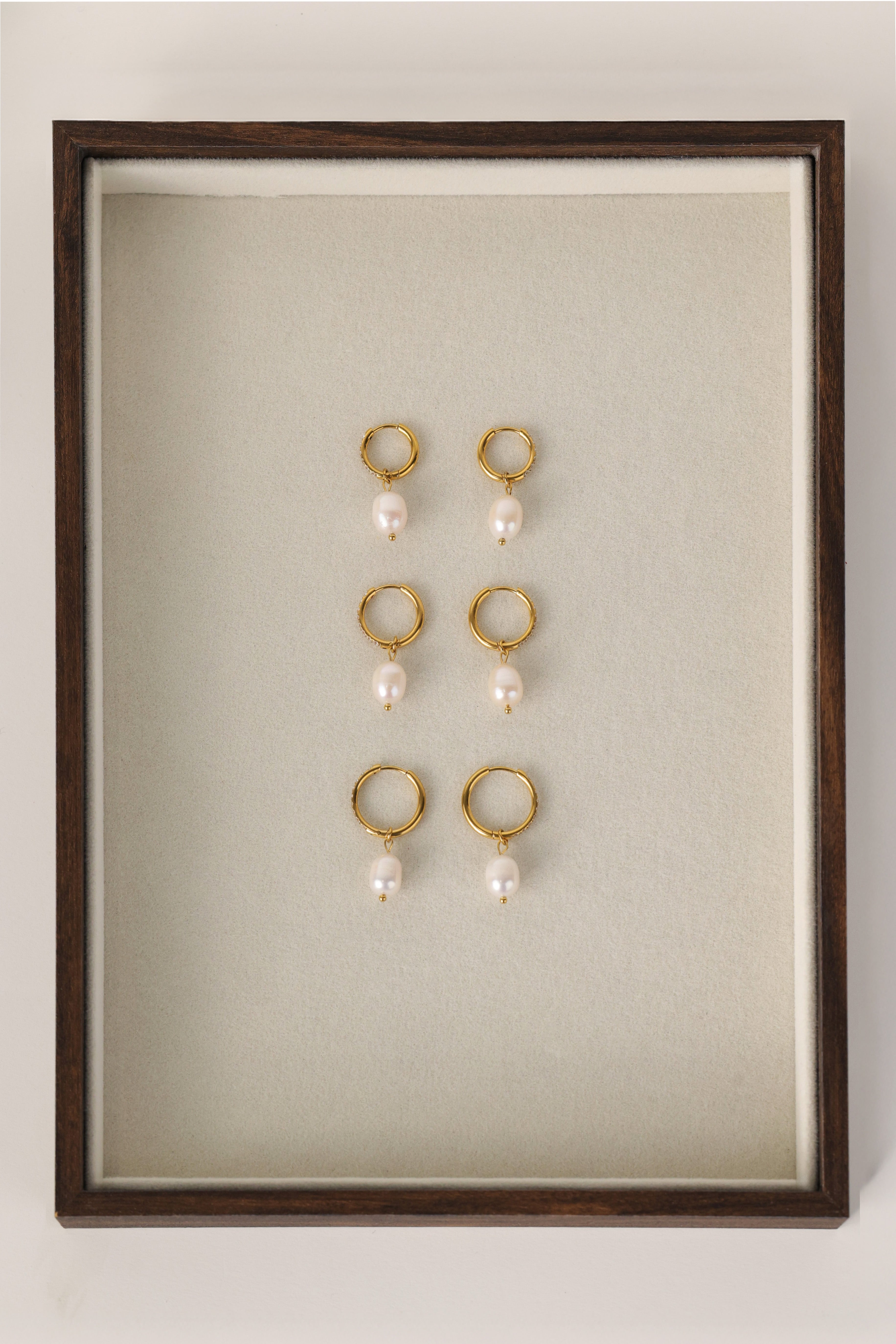 Three sizes of gold pavé waterproof hoop earrings with natural freshwater pearl drops displayed on a textured neutral background