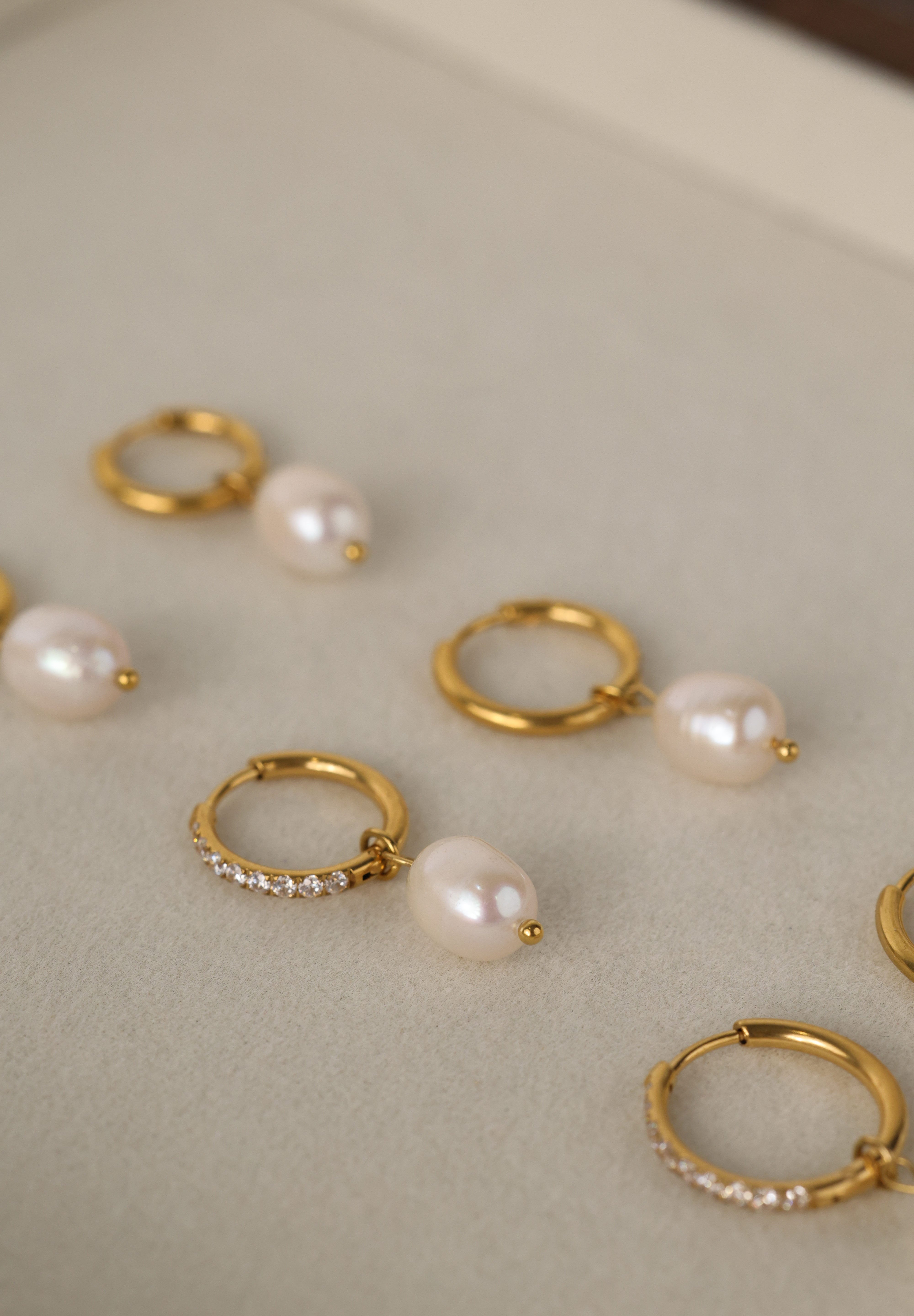 Three sizes of gold pavé waterproof hoop earrings with natural freshwater pearl drops displayed on a textured neutral background
