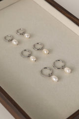 Six silver huggie hoop earrings, each adorned with a small cluster of clear pavé stones and a dangling white freshwater pearl, arranged on a beige jewelry display tray.