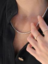 A close-up of a model wearing a gold tennis necklace with clear stones, styled with a black collared shirt and gold rings.