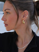 A close-up side profile of a woman wearing a gold huggie hoop earring and a delicate silver tennis necklace, with her hair in a ponytail.