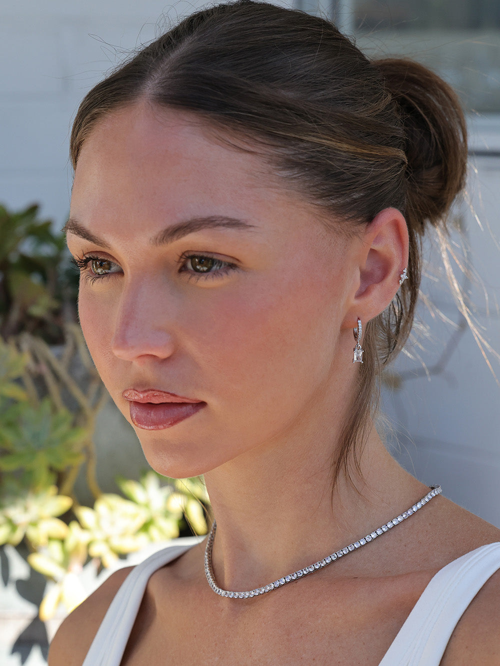 A close-up portrait of a woman wearing a delicate silver diamond necklace and matching drop earrings, with her hair in a bun.