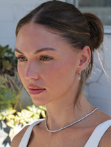 A close-up portrait of a woman wearing a delicate silver tennis necklace and matching drop earrings, with her hair in a bun.