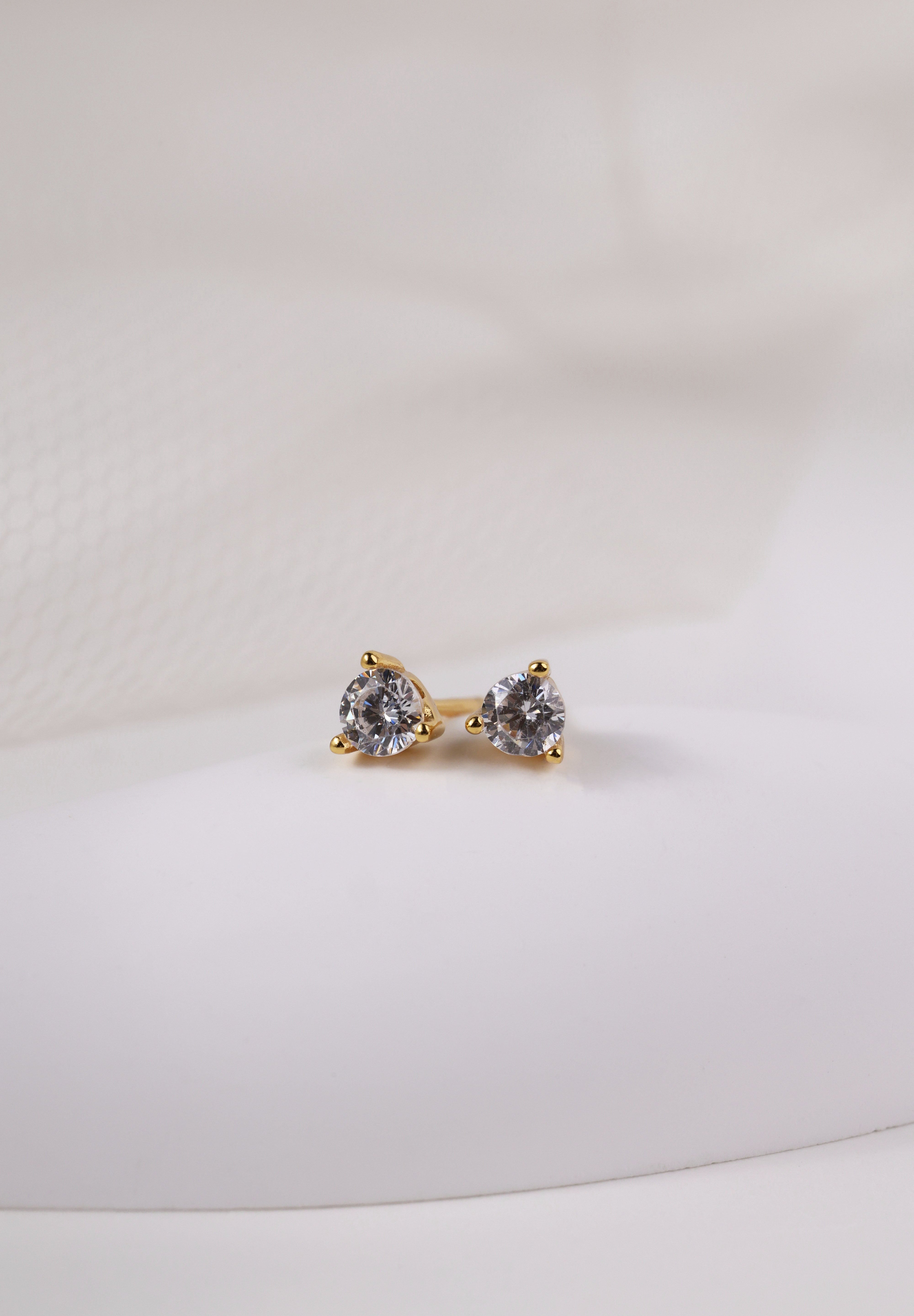 Pair of small gold round cubic zirconia stud earrings resting on cream fabric.