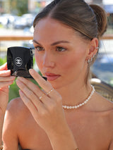 Woman holding a Chanel compact mirror outdoors, wearing silver earring stacks and pearl chocker necklace