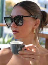 Side‑profile close‑up of a woman wearing gold hoop earrings, layered gold chain necklaces including one with a small pearl pendant, and a thin gold ring. She is outdoors with soft natural sunlight and blurred greenery in the background, showcasing an elegant everyday jewellery look.