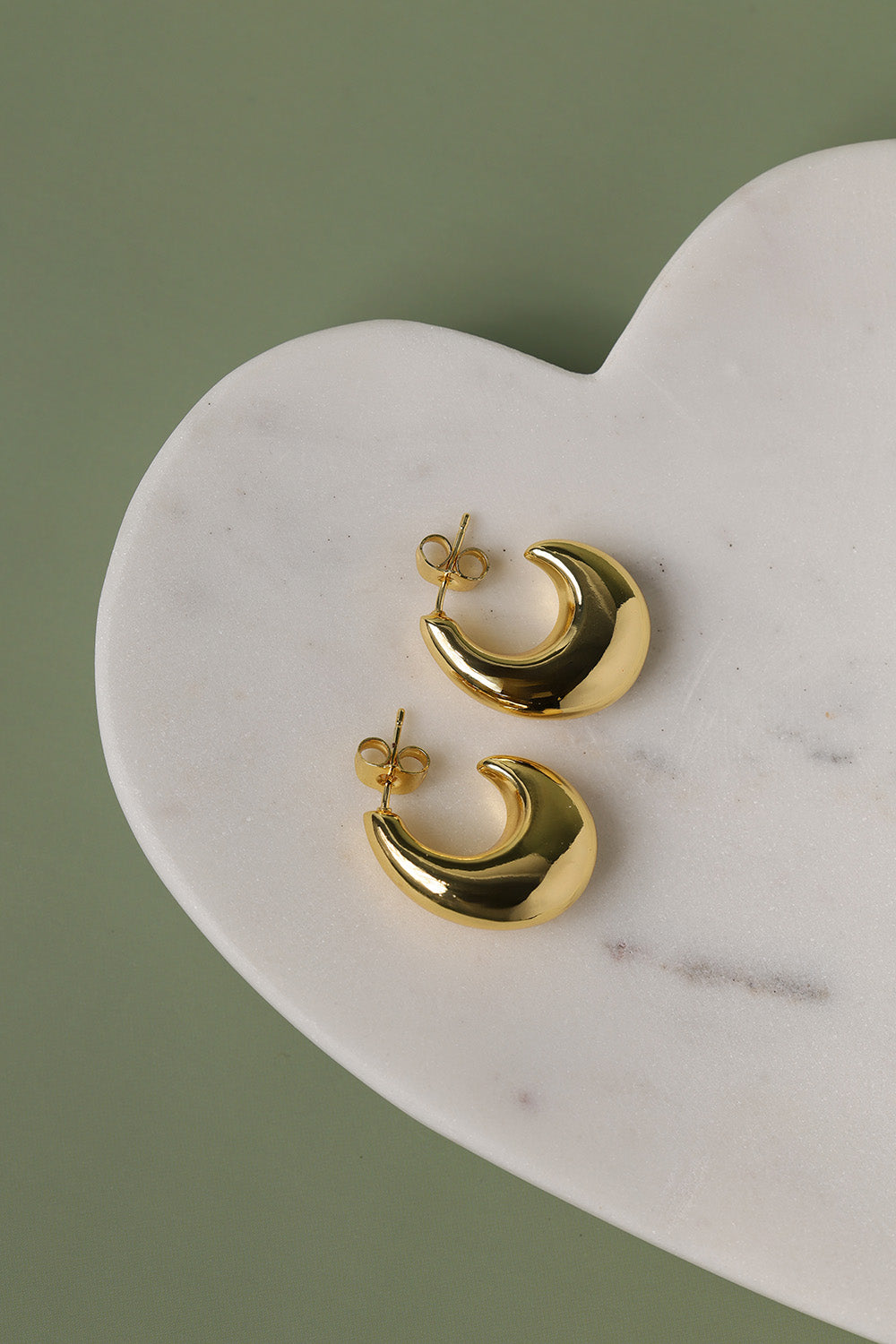 Pair of polished gold dome hoop earrings displayed on a heart-shaped marble tray against a sage green background