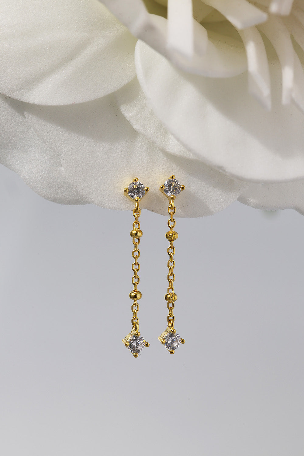 A pair of gold dangle earrings with clear gemstones, displayed against a soft white flower and a light gray background.