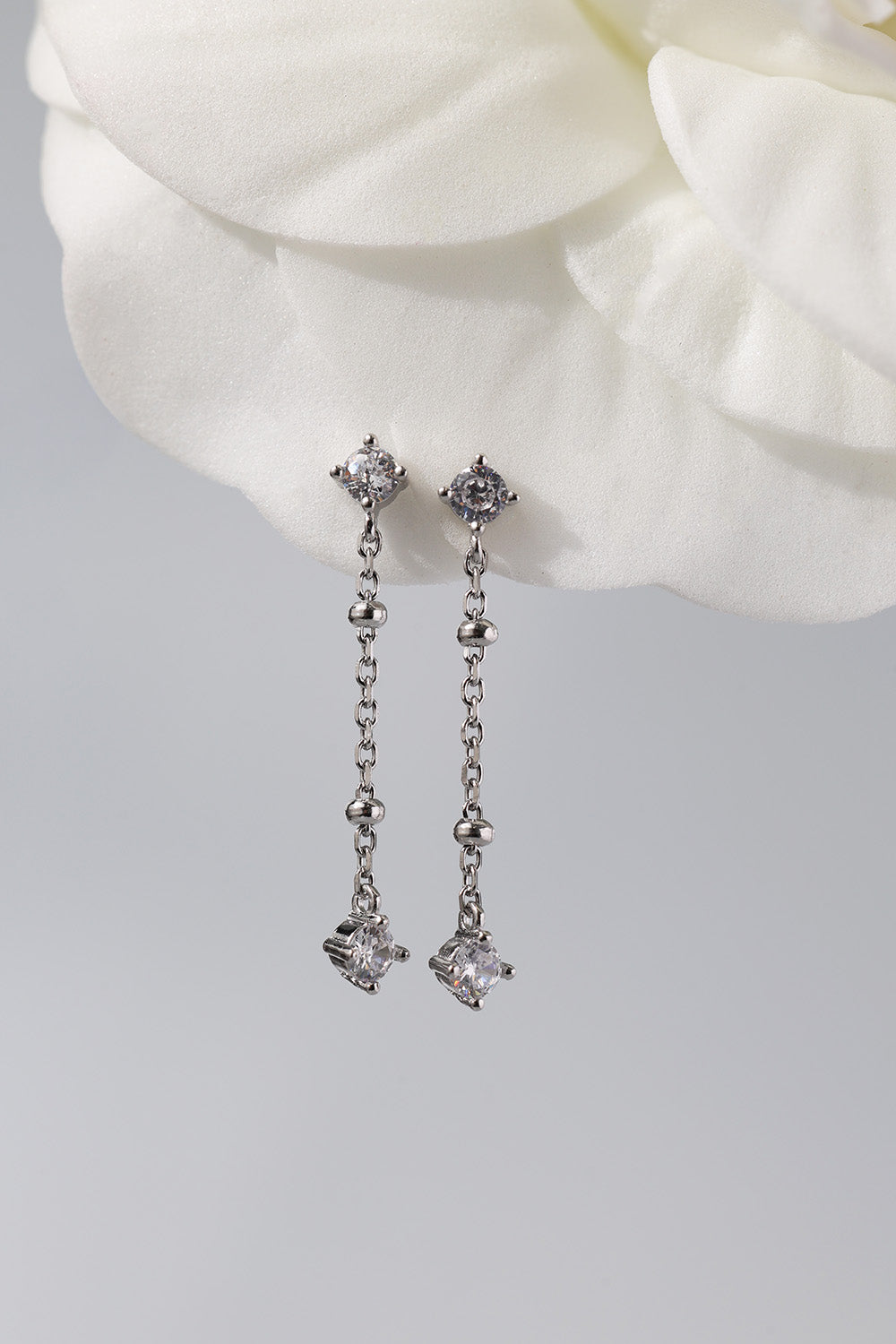 A pair of silver-toned dangle earrings with clear gemstones, displayed against a soft white flower and light gray background.