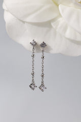 A pair of silver-toned dangle earrings with clear gemstones, displayed against a soft white flower and light gray background.