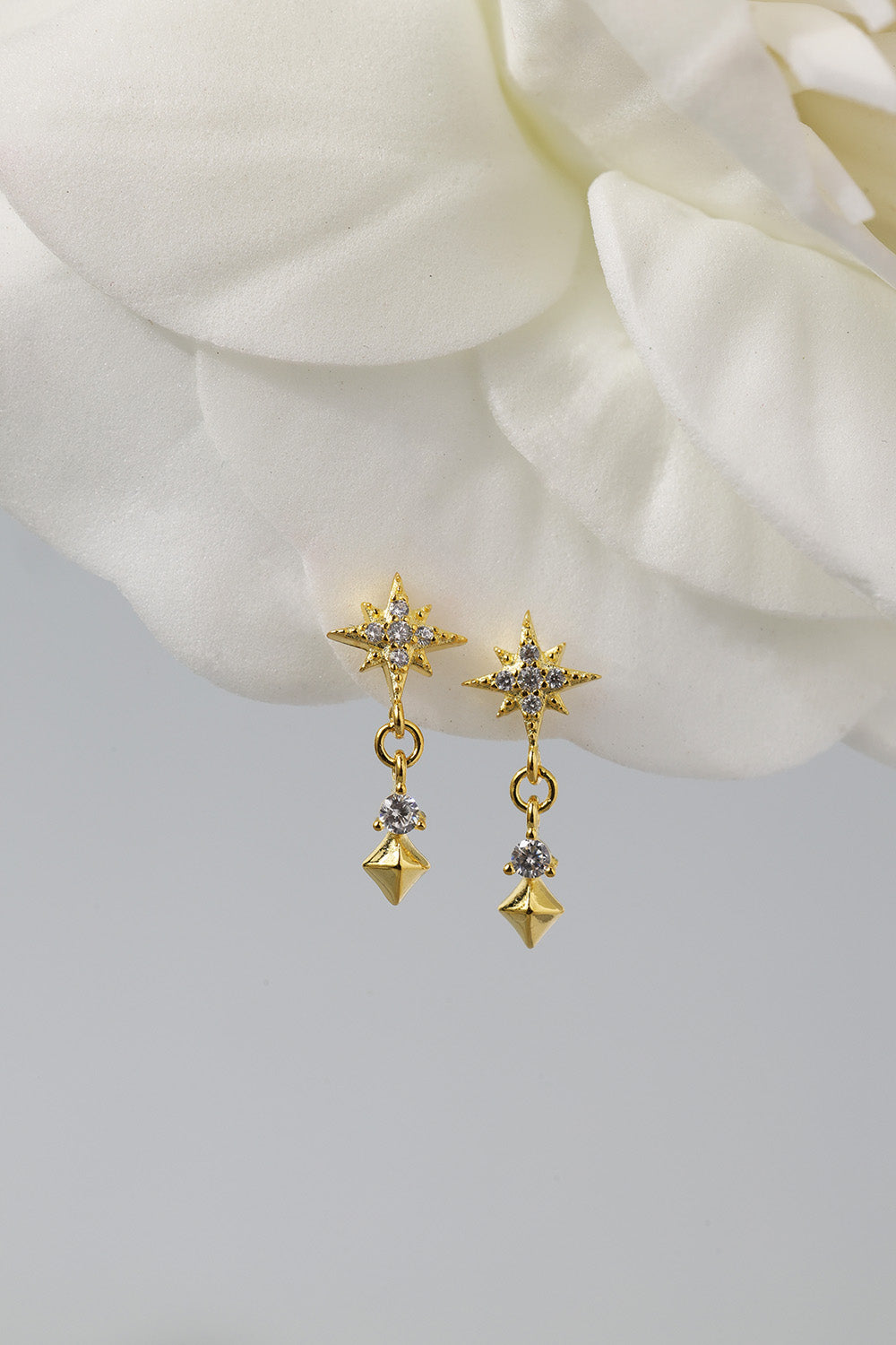 A pair of gold star-shaped dangle earrings with small clear stones and dangling pyramid charms, displayed against a soft white flower and light gray background.