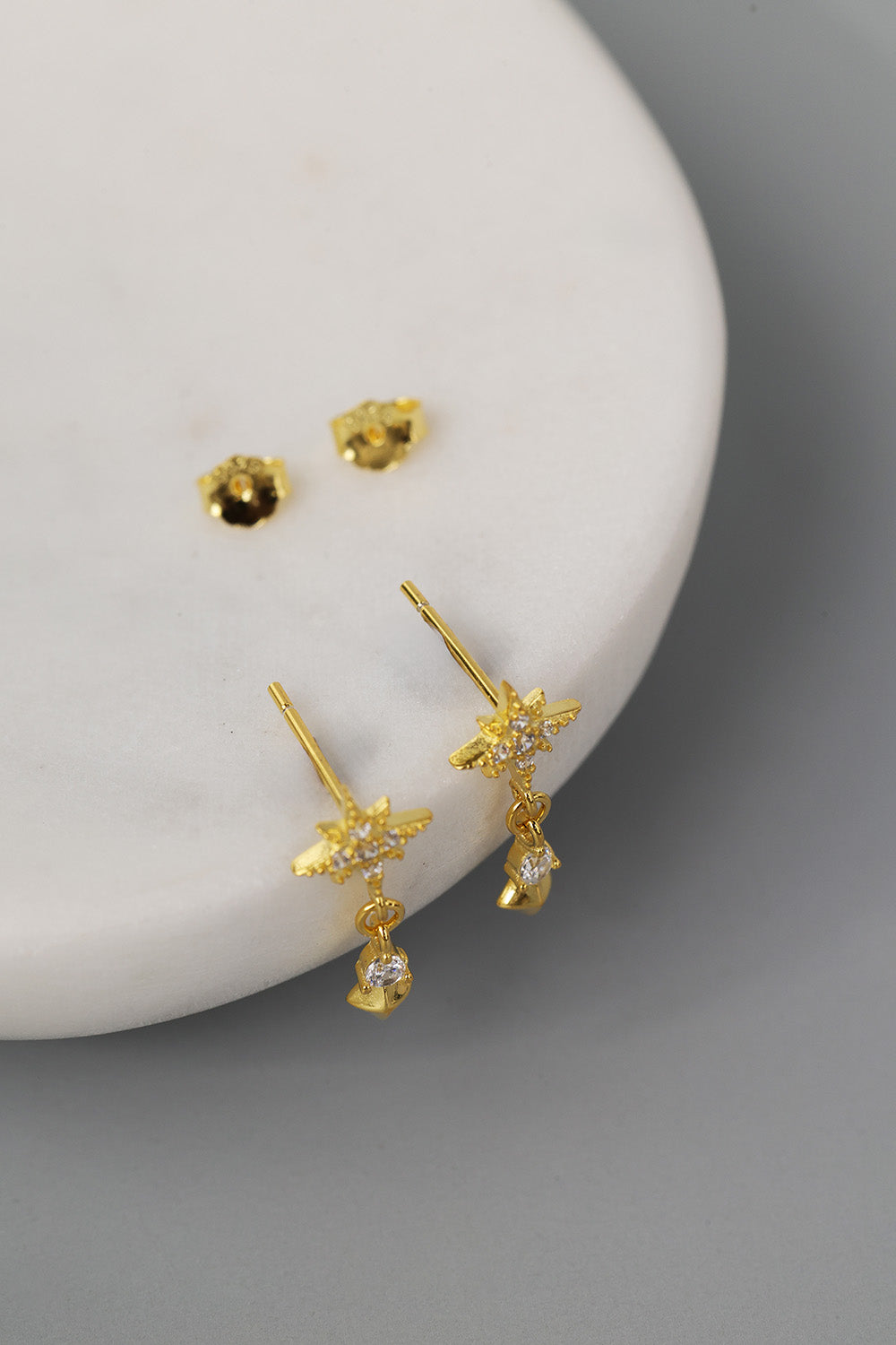 A pair of gold dangle earrings with star-shaped tops and small clear teardrop stones, displayed on a white curved surface against a gray background.