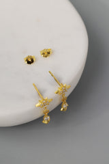 A pair of gold dangle earrings with star-shaped tops and small clear teardrop stones, displayed on a white curved surface against a gray background.