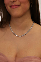 A close-up of a woman wearing a delicate silver tennis necklace with clear round-cut stones, paired with a soft pink strapless top.