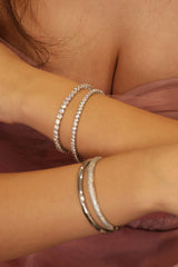 Close-up of a person's wrists wearing multiple silver bangles, including two tennis bracelets, against a soft mauve fabric background.