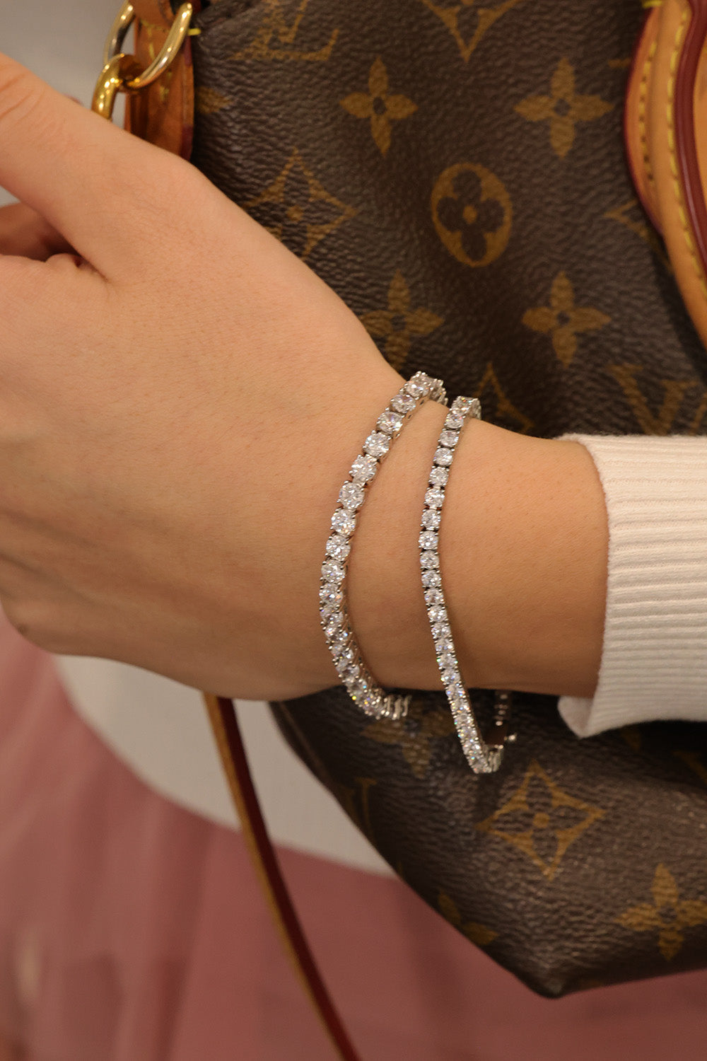 A close-up of a person's wrist wearing two silver tennis bracelets set with clear stones, next to a brown Louis Vuitton monogram handbag.