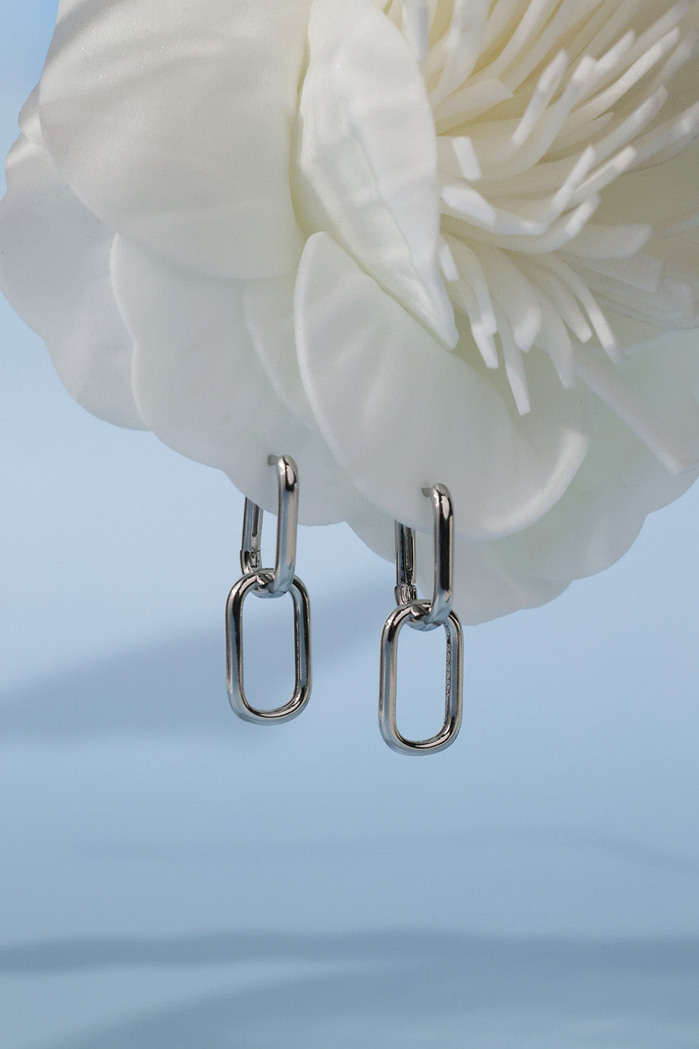 A pair of silver-toned drop earrings with elongated oval links, hanging in front of a large white flower against a soft light blue background.