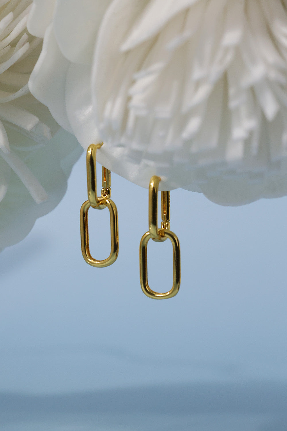 A pair of gold chain-link drop earrings hanging against a soft light blue background with a white flower prop at the top.