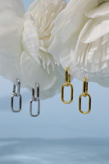 Two pairs of minimalist elongated oval drop earrings, one in polished silver and one in polished yellow gold, displayed against a soft light blue background with large white flower petals.
