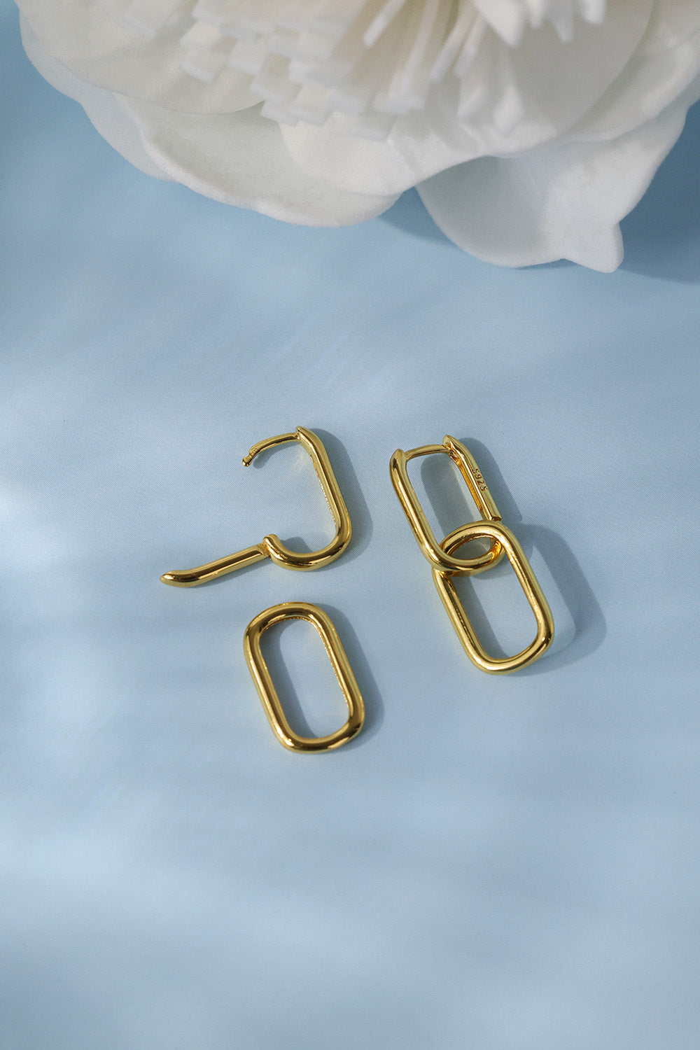 Gold geometric drop earrings with a chunky open oval and hook design, displayed on a light blue background with a white flower in the corner.
