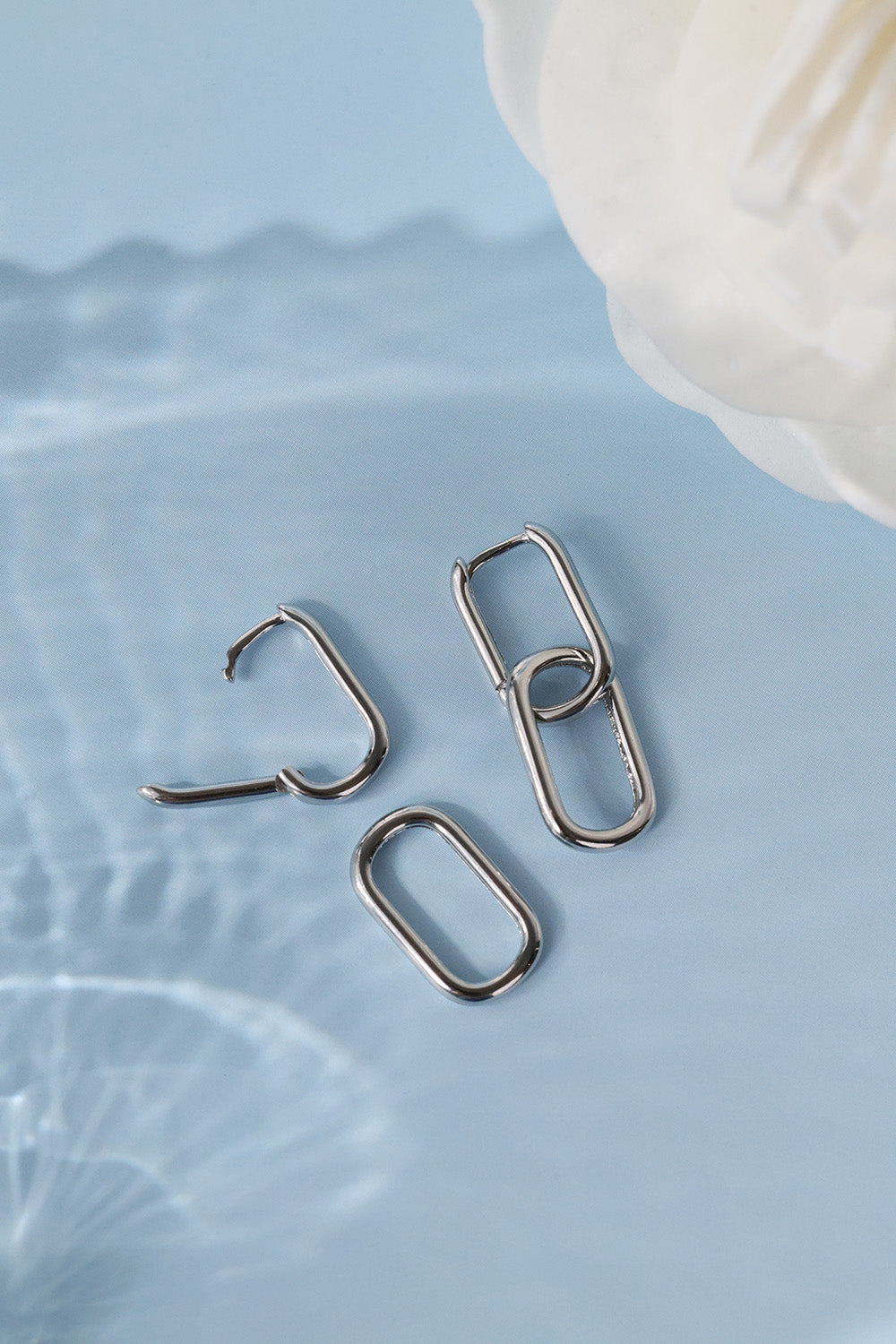 double silver metal ear cuffs with a minimalist open-link design, arranged on a light blue textured surface next to a white flower.