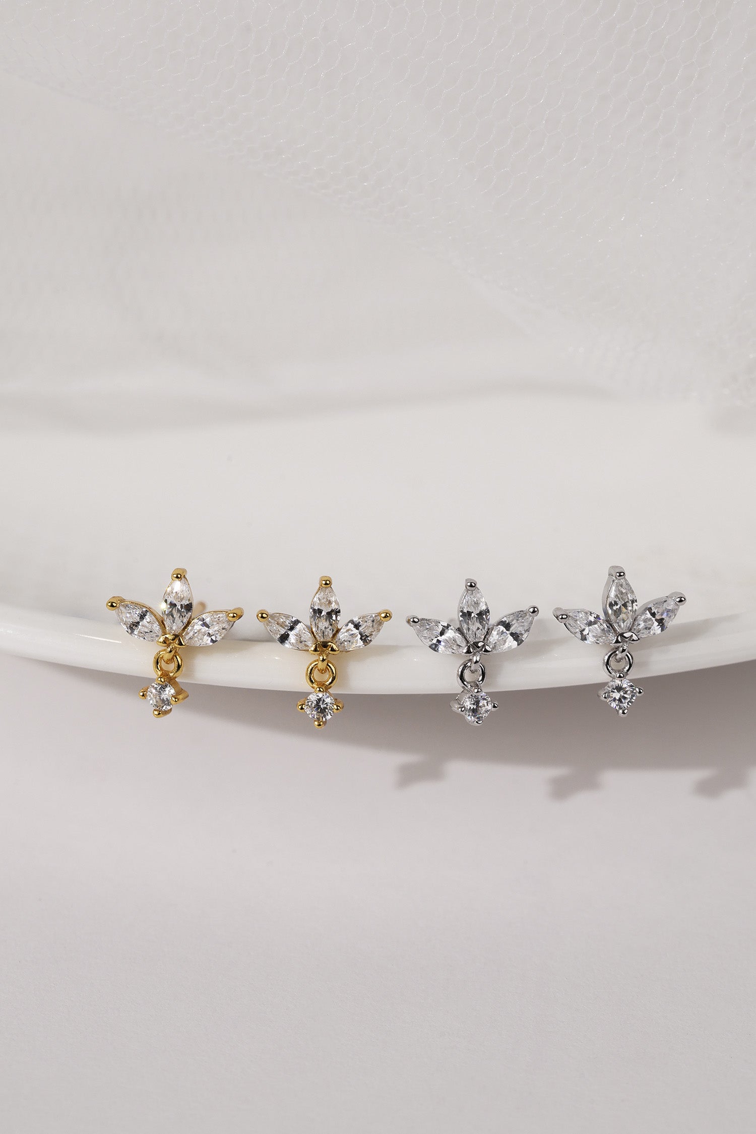 Four small cross-shaped stud earrings displayed on a white curved stand against a soft white background. Two earrings are gold-toned with clear stones, and two are silver-toned with clear stones.