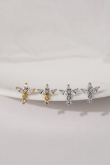 Four small cross-shaped stud earrings displayed on a white curved stand against a soft white background. Two earrings are gold-toned with clear stones, and two are silver-toned with clear stones.