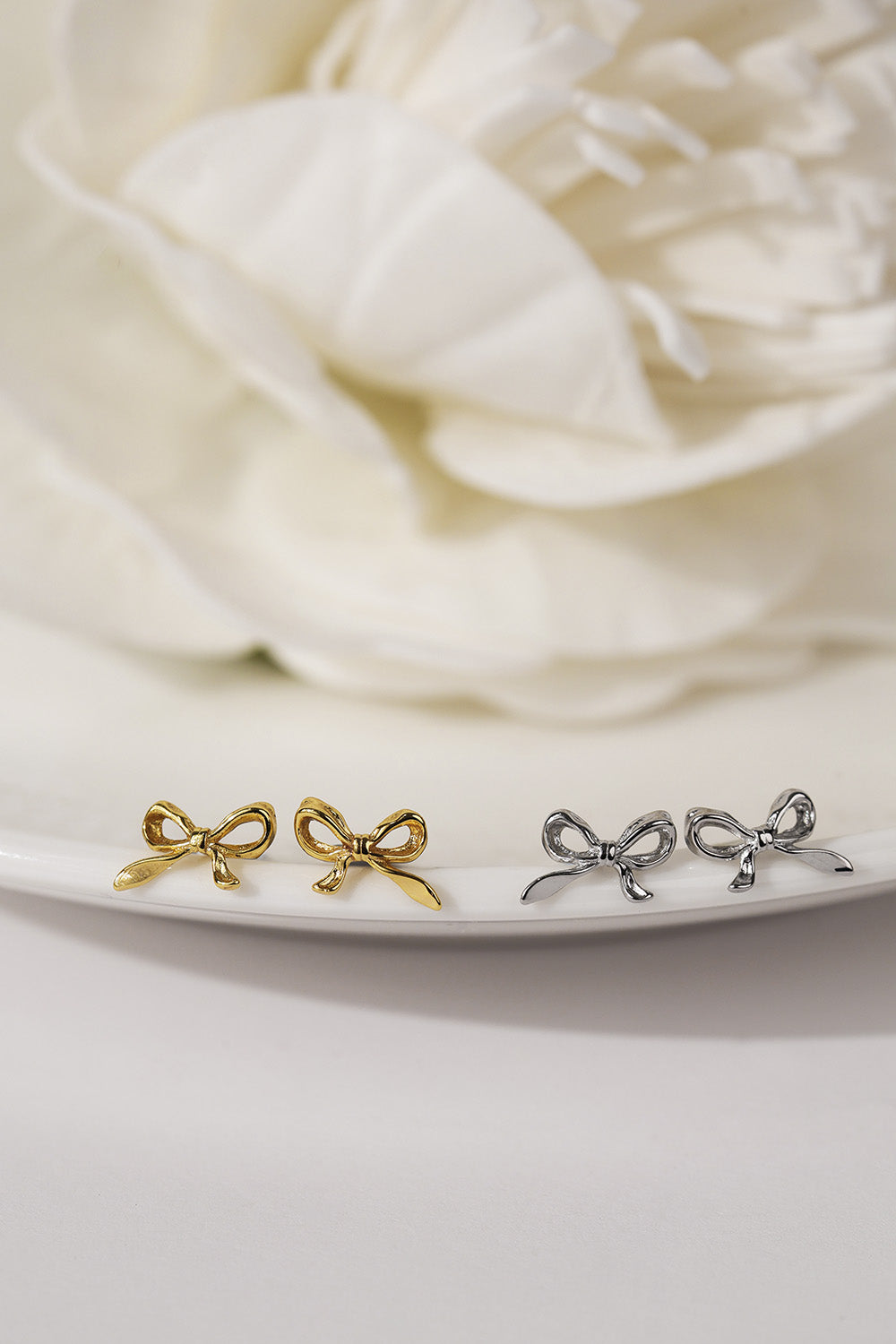 Two pairs of small bow-shaped stud earrings, one in polished yellow gold and one in polished silver, displayed on a white plate with a soft-focus white flower in the background.