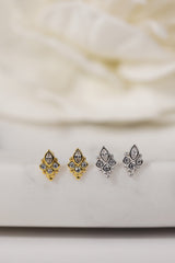 Three pairs of small stud earrings displayed on a light surface: two gold-tone pairs with clear stones and one silver-tone pair with clear stones, against a soft cream fabric background.