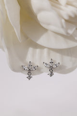 A pair of silver-toned dangle earrings with clear faceted gemstones, displayed on a soft white fabric surface with a blurred white flower in the background.