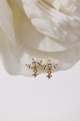 A pair of gold-toned dangle earrings with clear marquise and round crystals, displayed on the soft folds of a white rose.