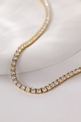 A close-up of a gold tennis necklace set with clear round-cut gemstones, resting on a soft white fabric surface.