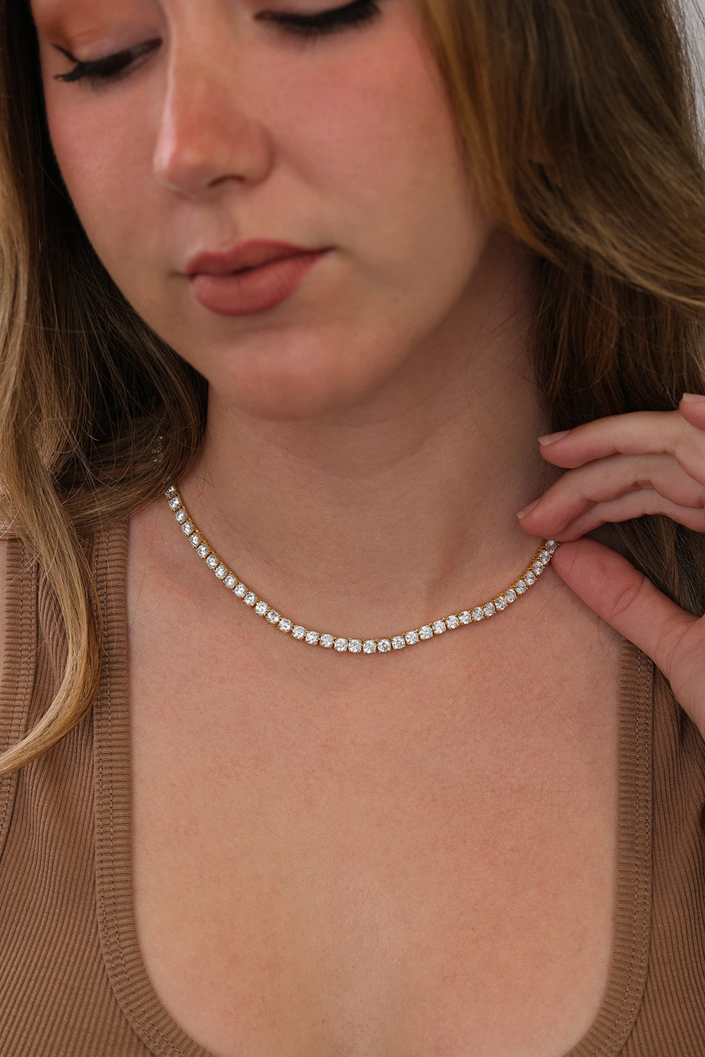 A close-up of a woman wearing a delicate gold tennis necklace with clear round-cut stones, paired with a brown ribbed top.