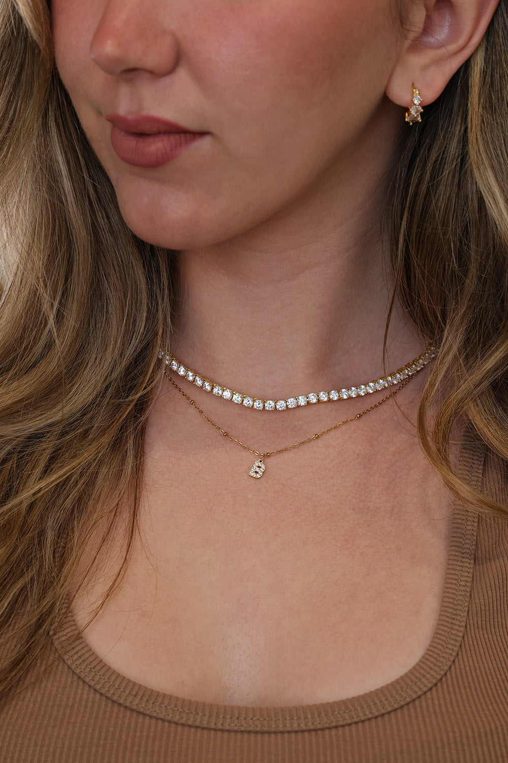 Close-up of a woman wearing a layered tennis necklace set with a short row of clear stones and a small gold initial pendant, along with a matching gold earring.