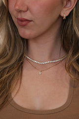 Close-up of a woman wearing a layered tennis necklace set with a short row of clear stones and a small gold initial pendant, along with a matching gold earring.