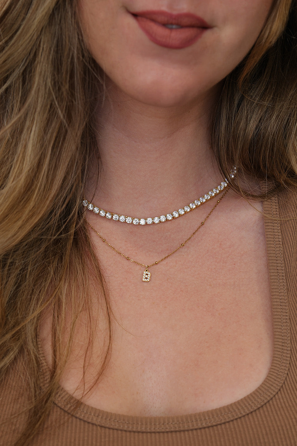 Close-up of a woman wearing a delicate gold 4mm tennis necklace with a small initial 'B' pendant and a thin rhinestone tennis-style choker.