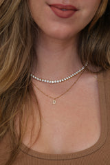 Close-up of a woman wearing a delicate gold 4mm tennis necklace with a small initial 'B' pendant and a thin rhinestone tennis-style choker.