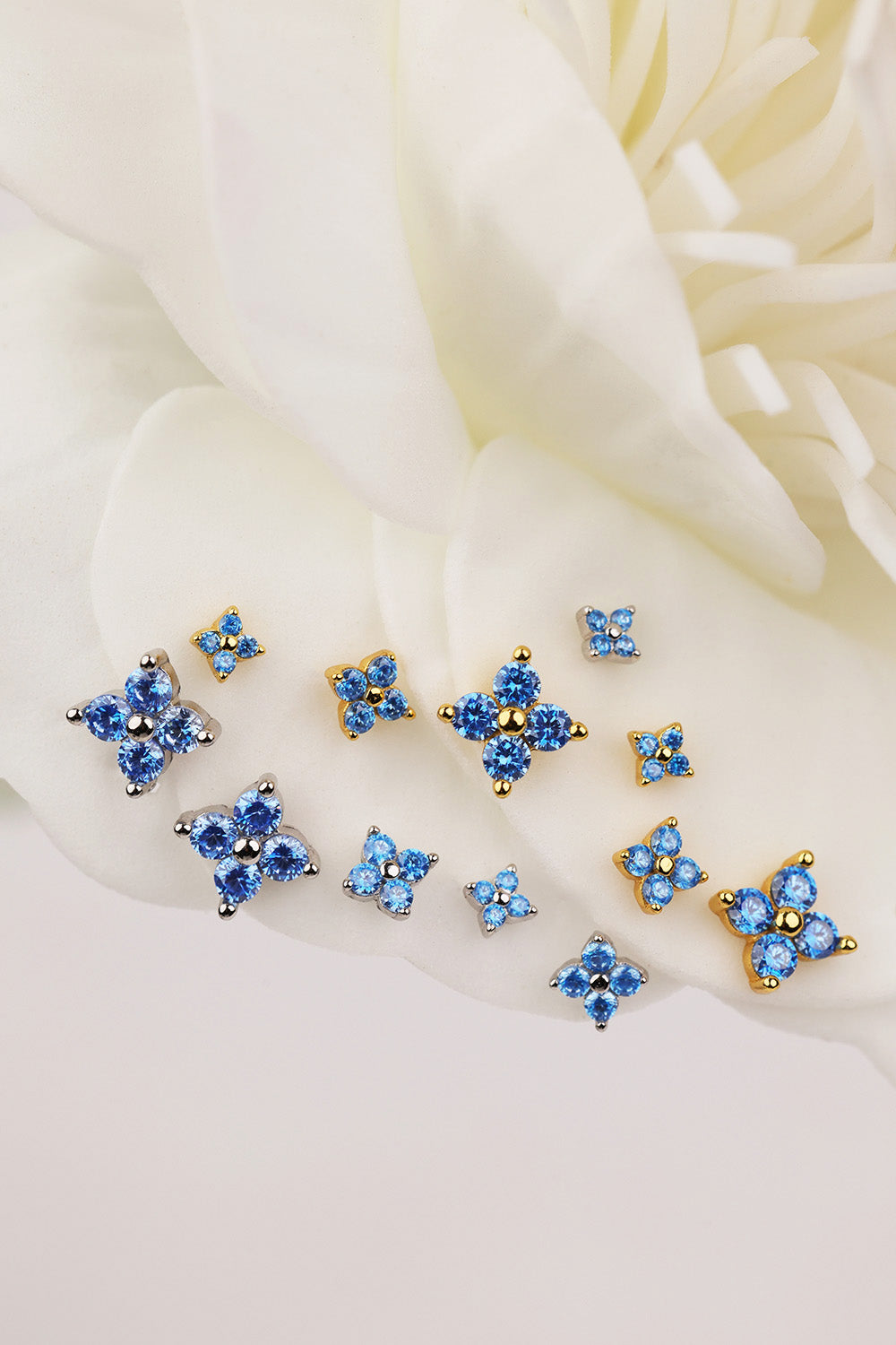 A set of small blue floral 4 clover stud earrings with gold-tone settings, arranged on soft white flower petals.