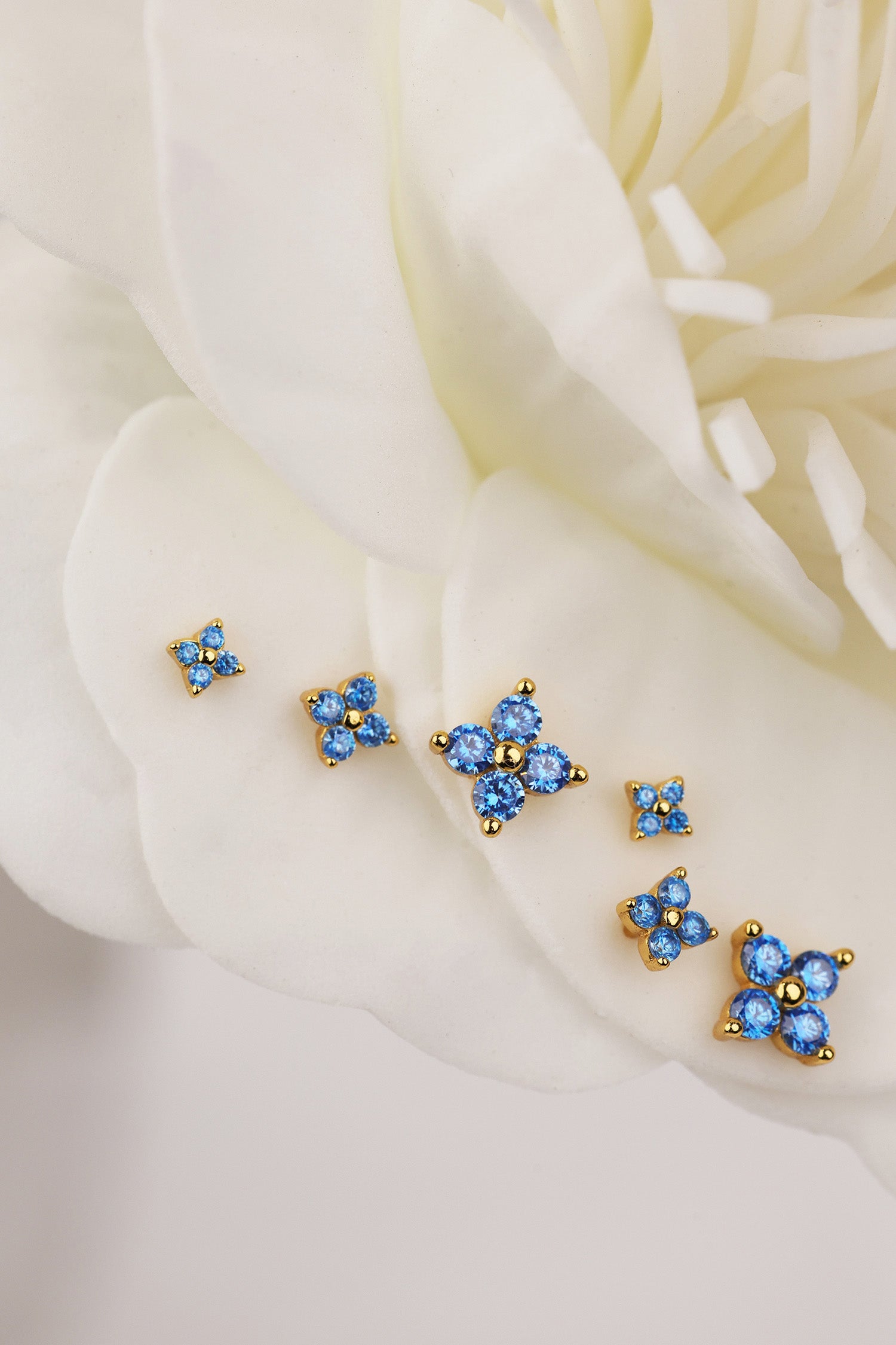 A set of small gold-tone 4 clover stud earrings with blue crystal clusters, arranged on soft white flower petals.
