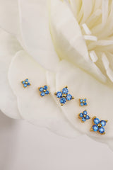 A set of small gold-tone 4 clover stud earrings with blue crystal clusters, arranged on soft white flower petals.