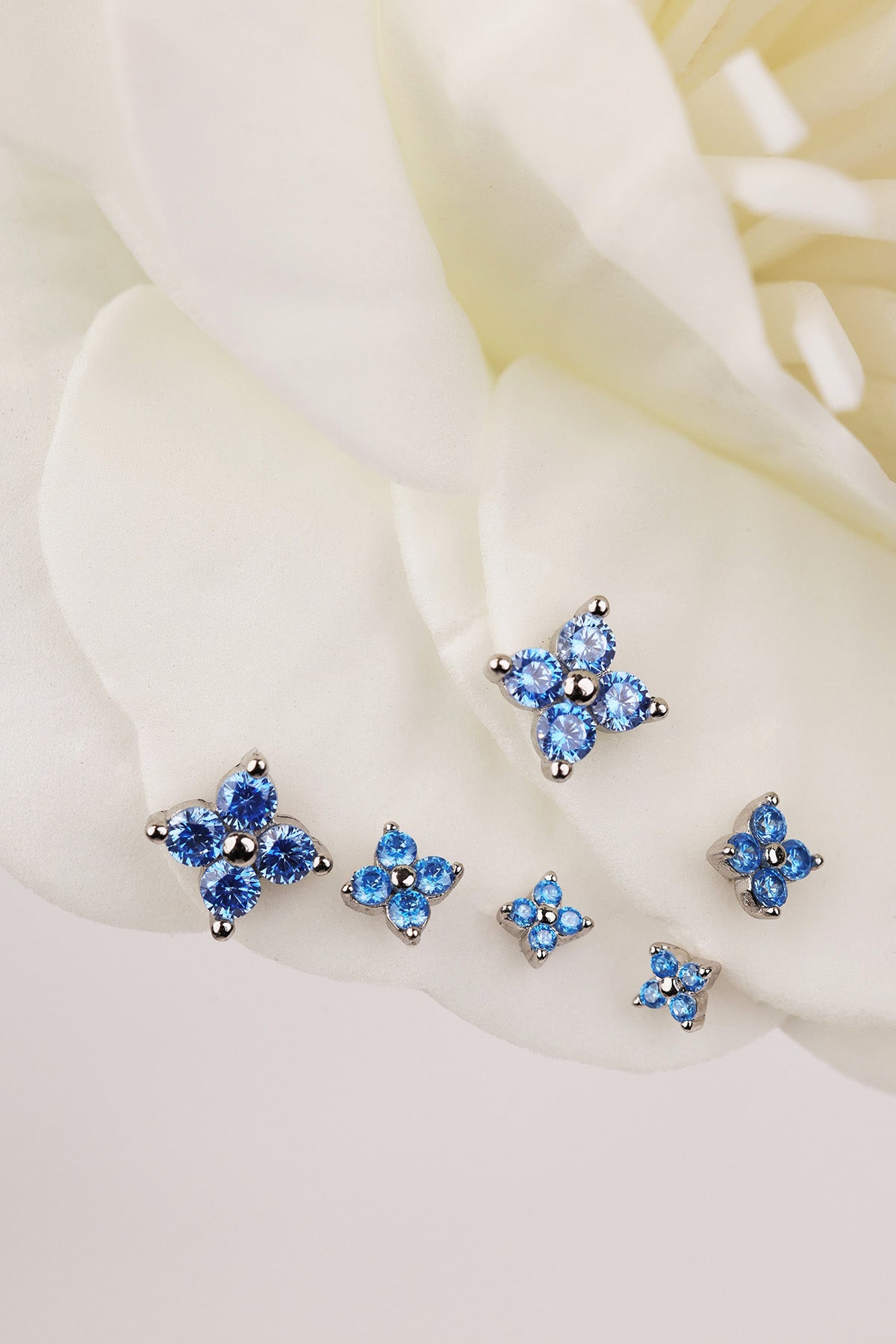 A set of five silver-toned 4 clover stud earrings with blue gemstone clusters, arranged on soft white flower petals.