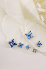A set of five silver-toned 4 clover stud earrings with blue gemstone clusters, arranged on soft white flower petals.