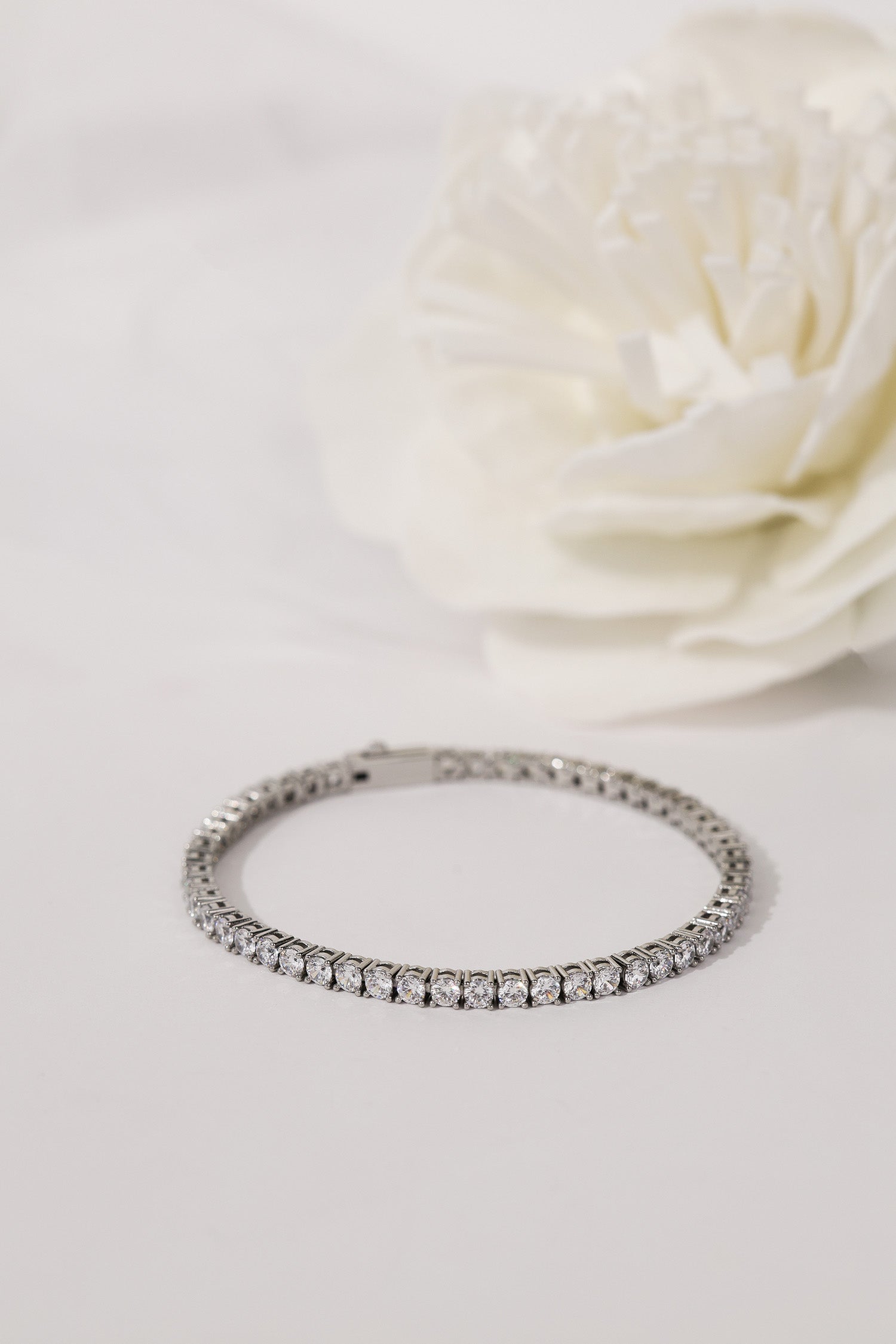 A silver tennis bracelet with a continuous row of clear gemstones, displayed on a white surface with a soft-focus white flower in the background.