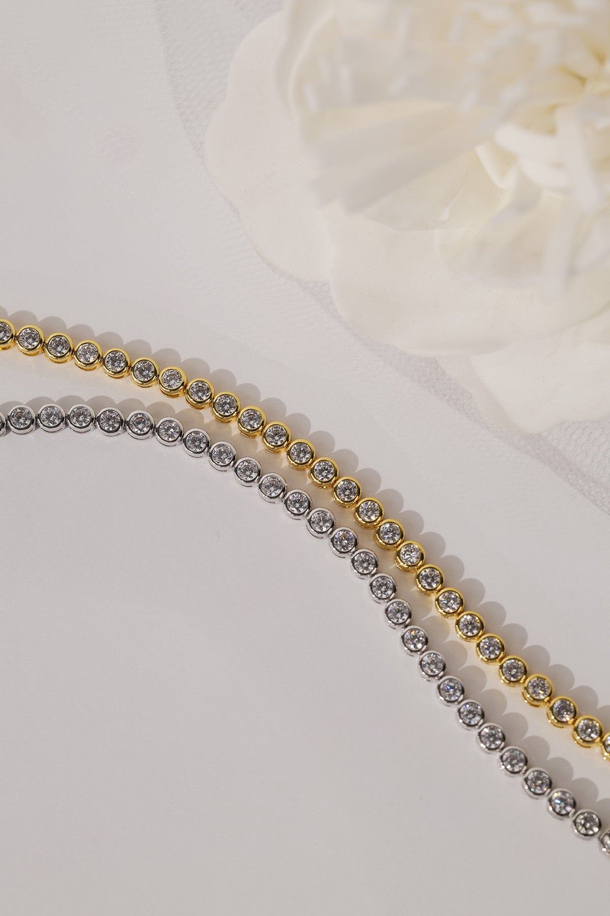 Two tennis-style bead chains laid diagonally on a light surface, one in yellow gold and one in silver, with a soft white flower prop in the background.