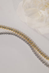 Two tennis-style bead chains laid diagonally on a light surface, one in yellow gold and one in silver, with a soft white flower prop in the background.